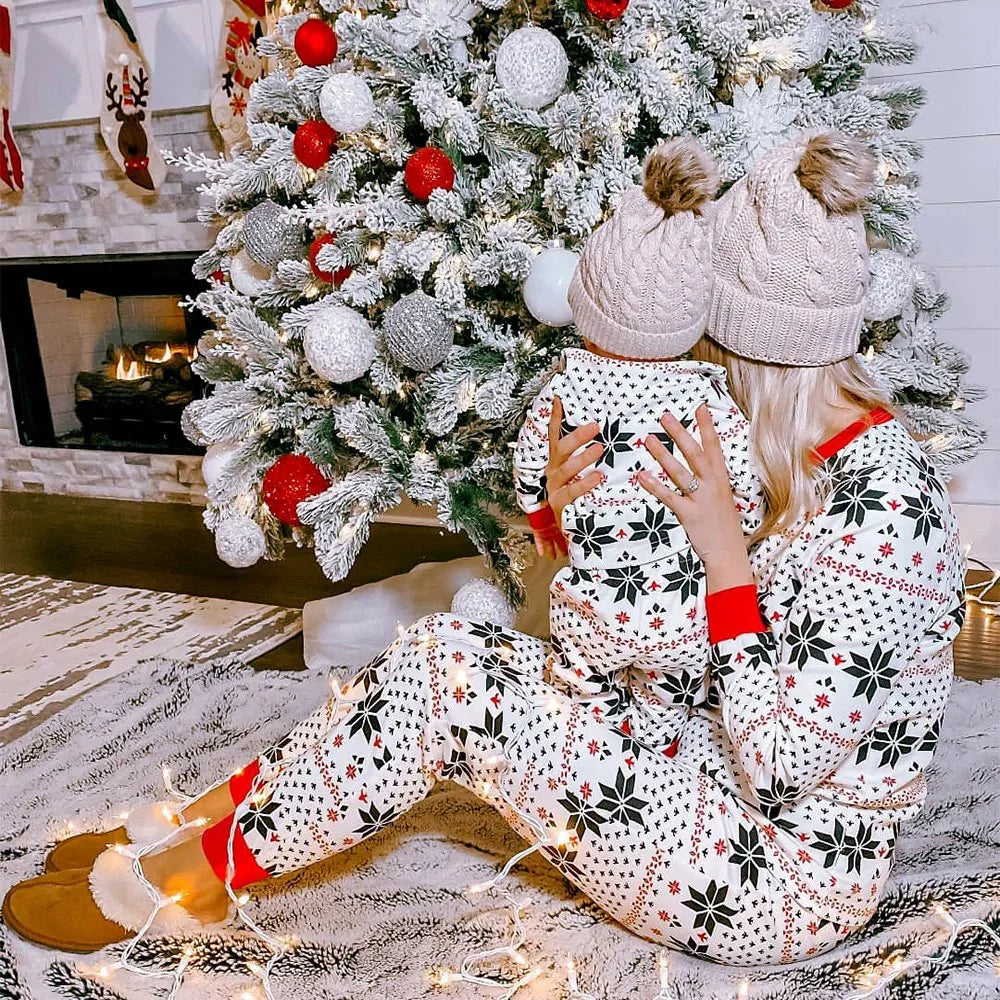 Mother and toddler by a snowy Christmas tree wearing matching white Fair Isle snowflake pajamas with knit beanies.