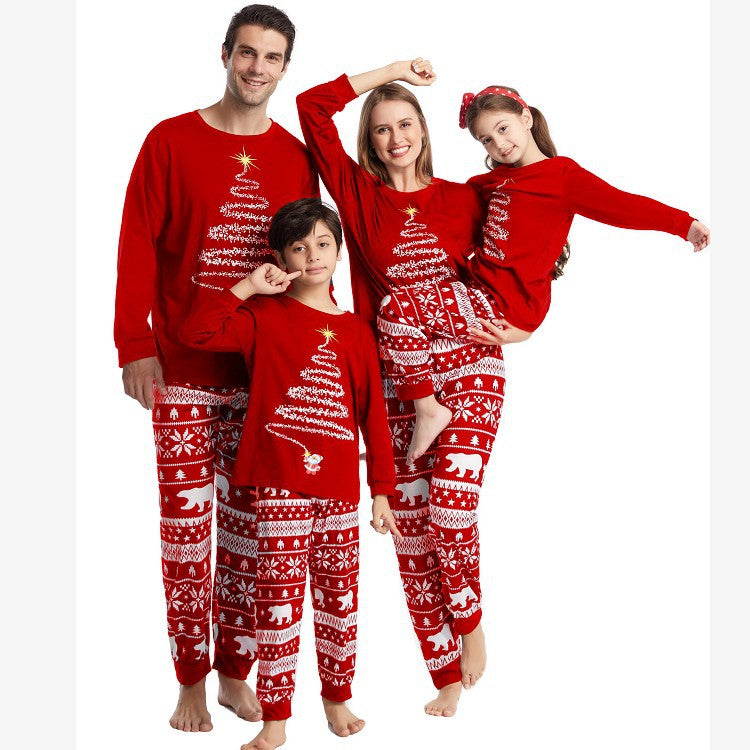 Full-body standing studio photo of a family in matching red holiday PJs, featuring mom holding her daughter in a playful pose wearing long-sleeve Christmas tree crewnecks and Fair Isle pants.