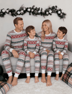 Parents and two boys smiling at each other on a sofa in grey pajamas with red reindeer and green Christmas trees.