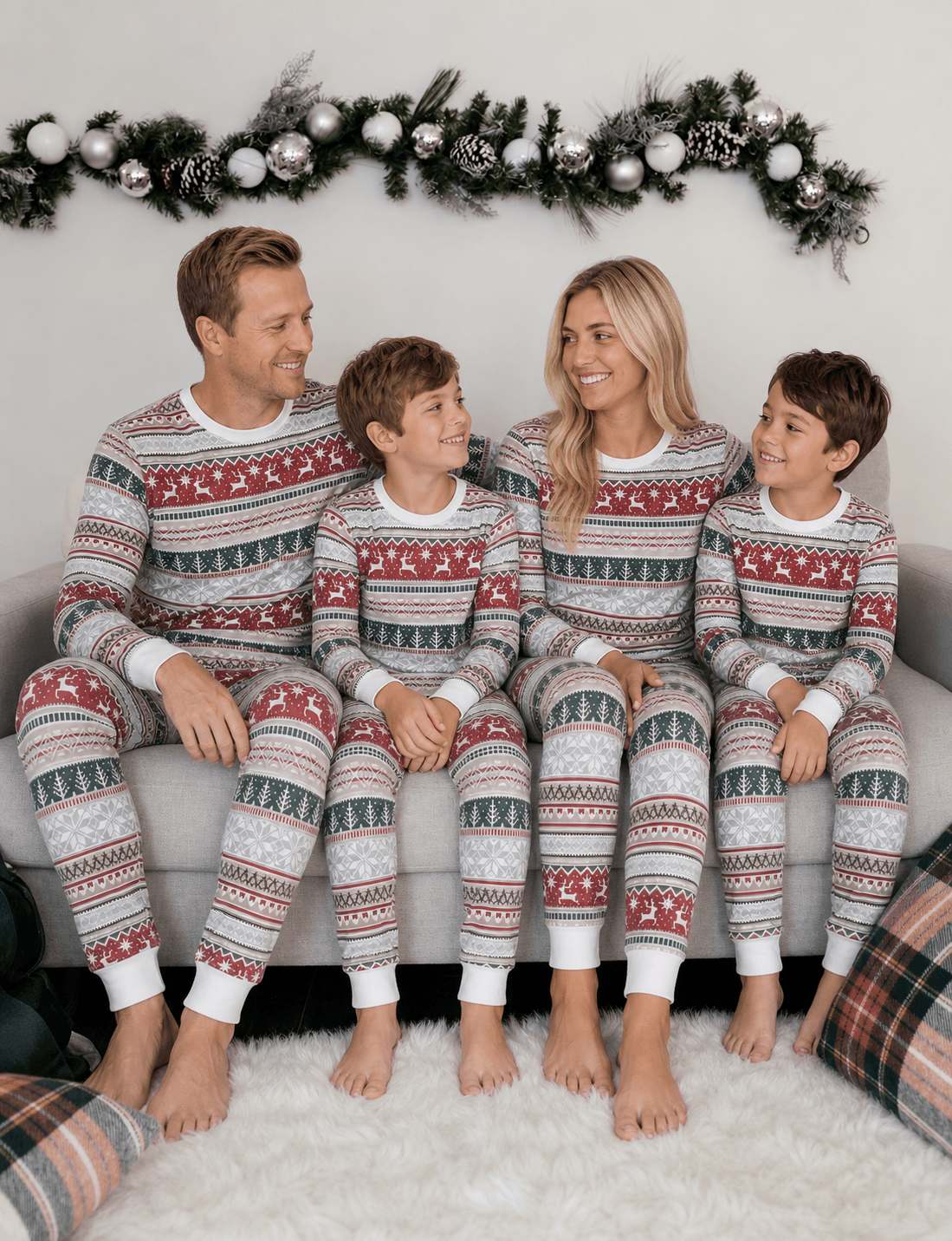 Parents and two boys smiling at each other on a sofa in grey pajamas with red reindeer and green Christmas trees.