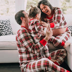 Pregnant mother laughing with her husband and toddler on a white couch, all wearing matching cream red plaid holiday loungewear.