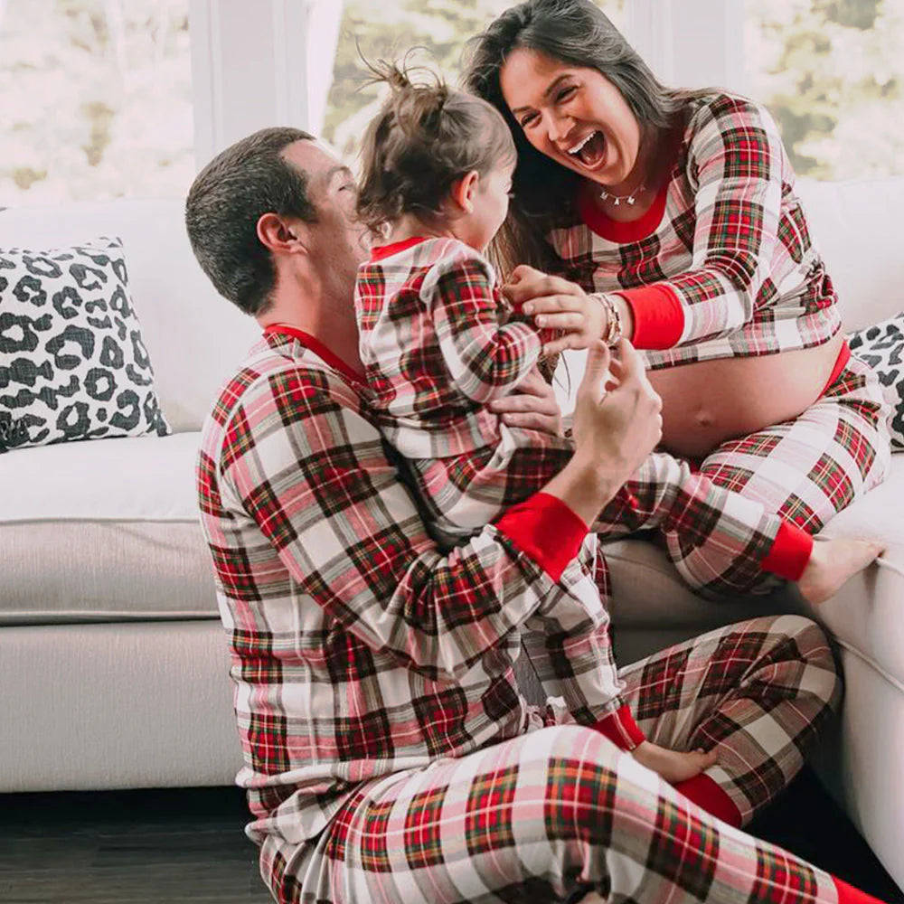 Pregnant mother laughing with her husband and toddler on a white couch, all wearing matching cream red plaid holiday loungewear.