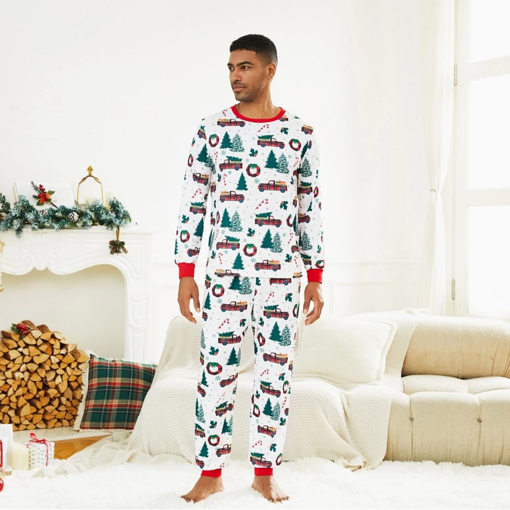 Adult man modeling a white two-piece holiday pajama set with red truck and Christmas tree print in a bright living room.