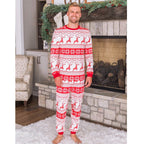 Smiling adult man modeling a men's red and white Fair Isle reindeer Christmas pajama set, standing front view on a white rug in front of a decorated stone fireplace.