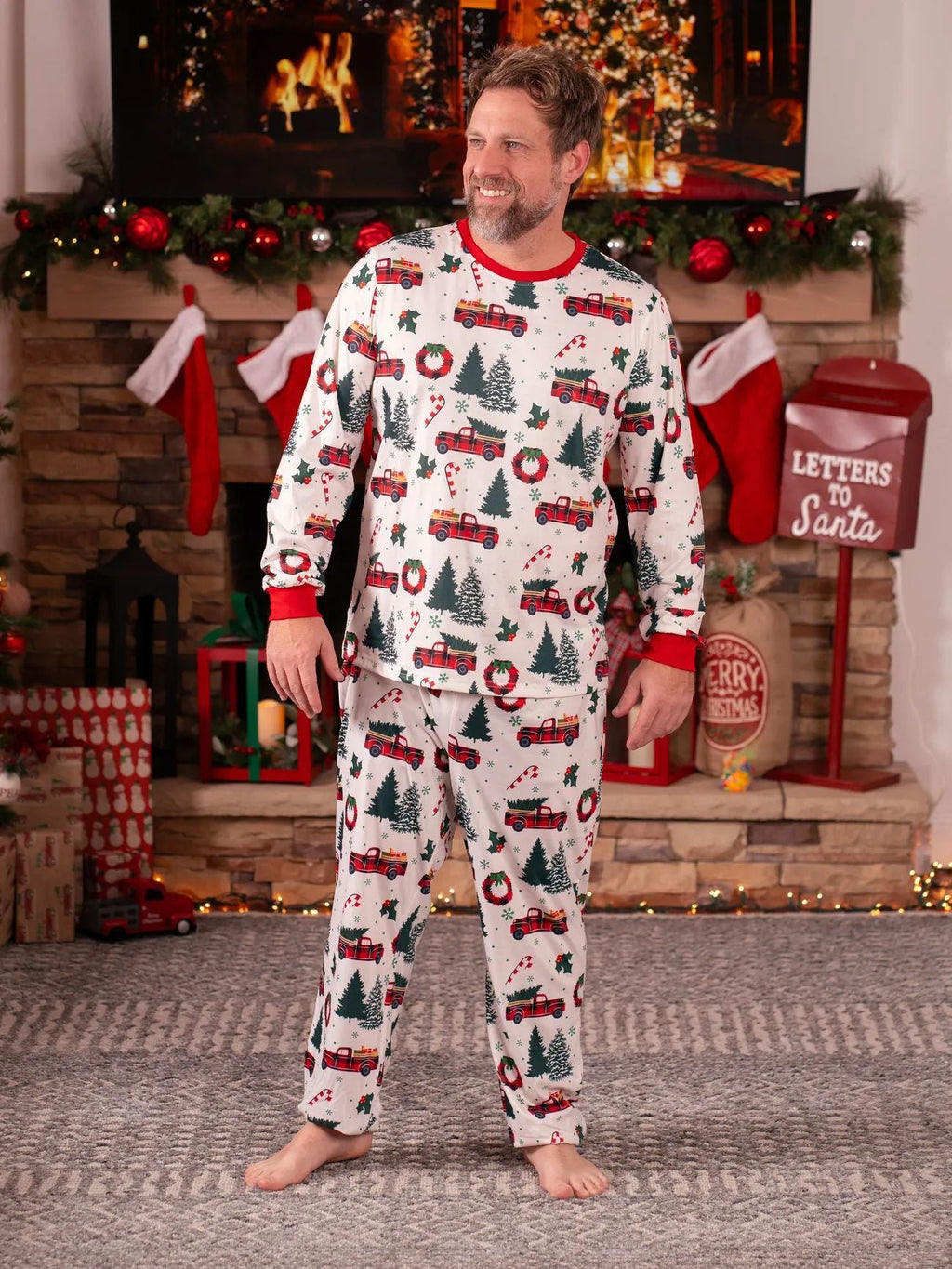 Smiling man wearing a white long-sleeve Christmas pajama set with vintage red truck and tree patterns by a fireplace.