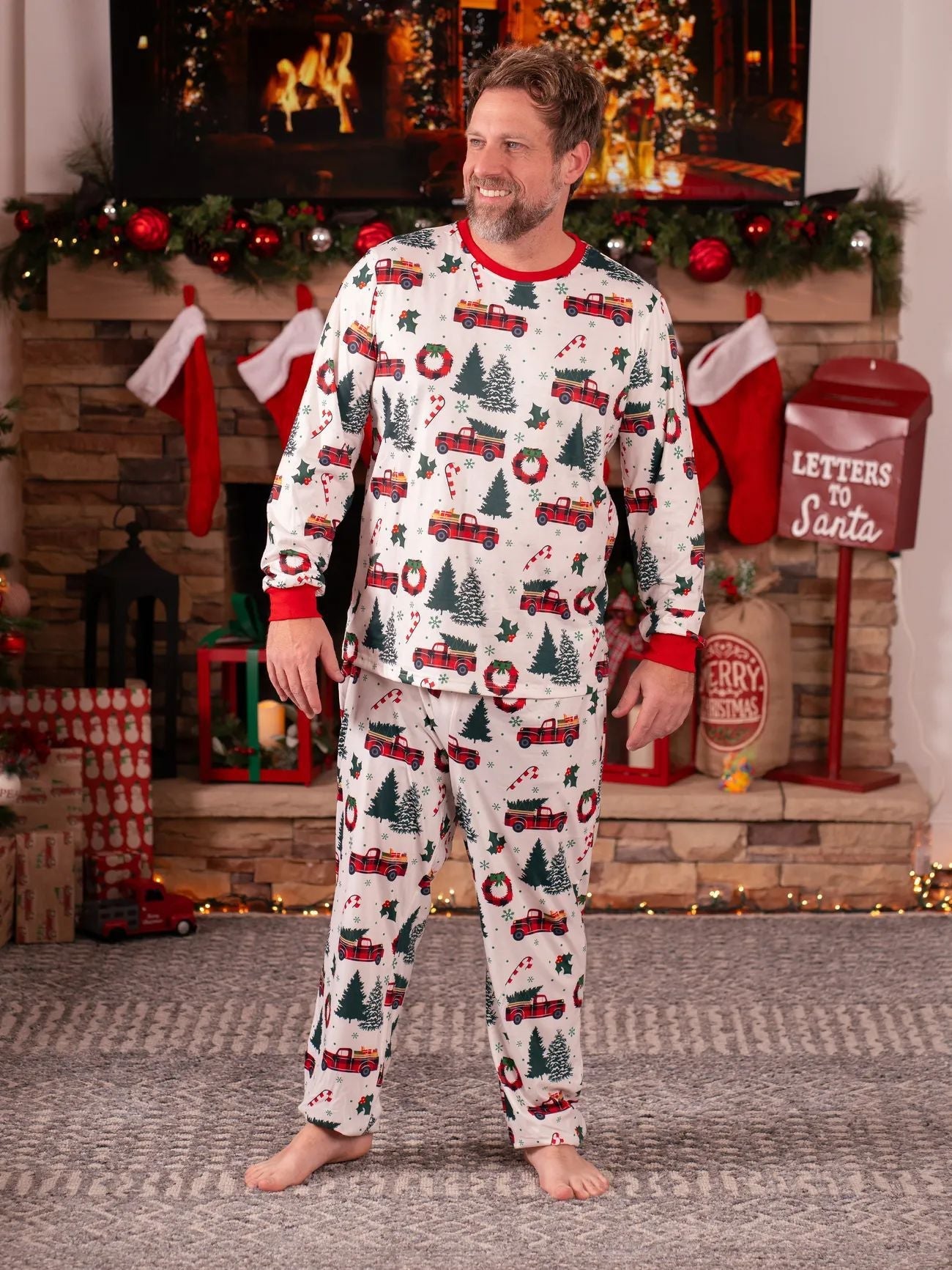 Smiling man wearing a white long-sleeve Christmas pajama set with vintage red truck and tree patterns by a fireplace.