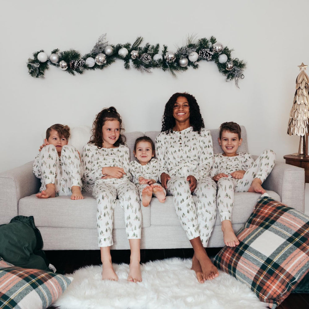 A mother sitting on a neutral sofa surrounded by her four children, all wearing matching white holiday loungewear printed with green watercolor pine trees under evergreen garland.