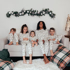 A mother sitting on a neutral sofa surrounded by her four children, all wearing matching white holiday loungewear printed with green watercolor pine trees under evergreen garland.