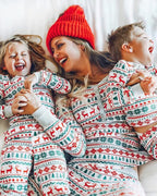 Laughing mother wearing a red beanie lying on a bed with her two children in matching white, red, and green Fair Isle reindeer Christmas pajamas.