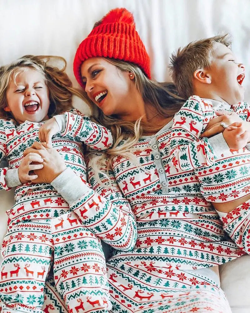 Laughing mother wearing a red beanie lying on a bed with her two children in matching white, red, and green Fair Isle reindeer Christmas pajamas.