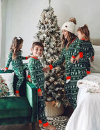 Mother wearing a white beanie holding her toddler next to two kids playing near a flocked Christmas tree in matching green Fair Isle pajamas.