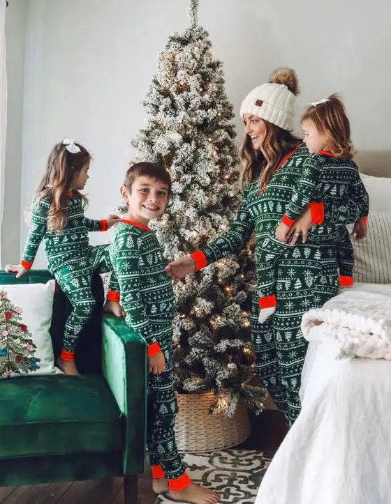 Mother wearing a white beanie holding her toddler next to two kids playing near a flocked Christmas tree in matching green Fair Isle pajamas.