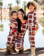 Mother hugging her two young sons outdoors near a lake wearing matching cream, red, and green tartan plaid Christmas pajamas.
