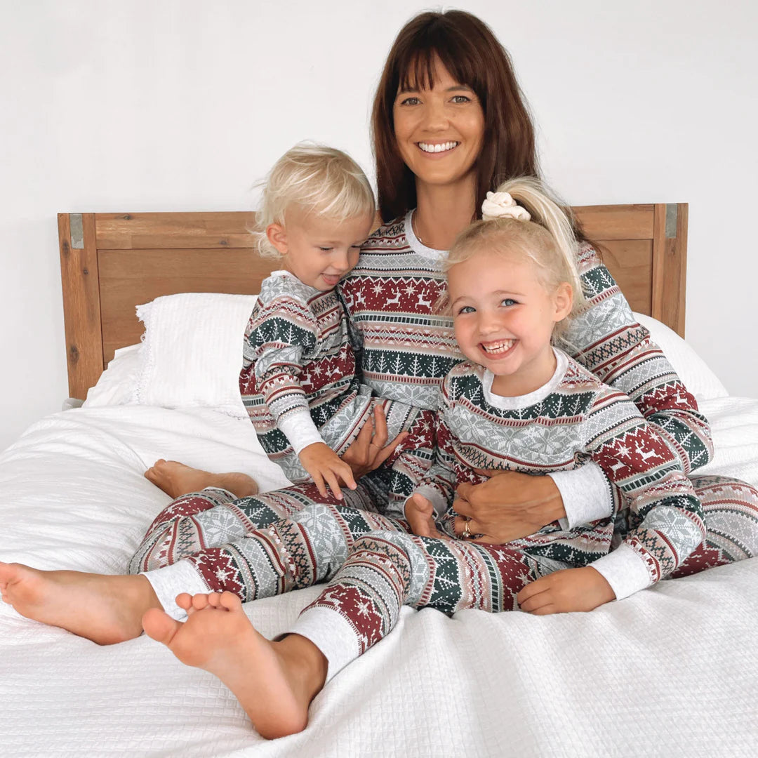 Happy mother holding her two young toddlers on a white bed, all wearing matching grey and burgundy Fair Isle reindeer print holiday pajama sets.