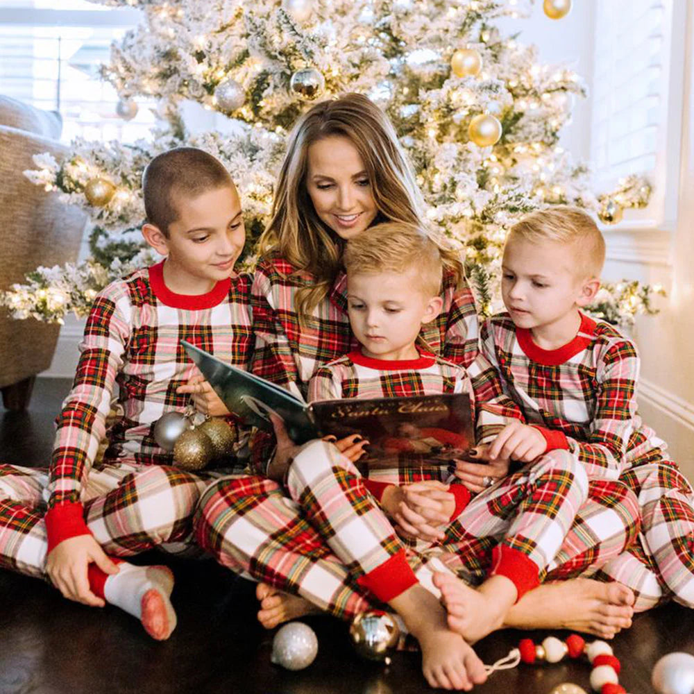 Mother reading a Christmas storybook to her three young sons sitting under a lighted frosted Christmas tree in matching cream, red, and green plaid pajama sets.
