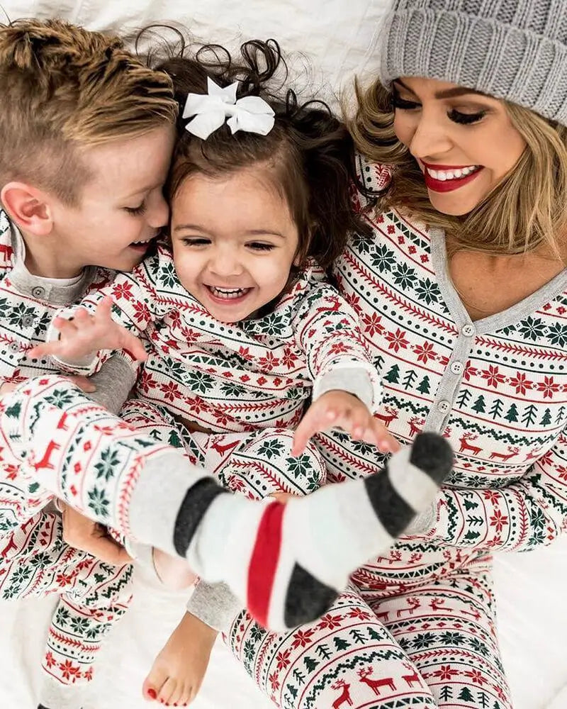 Mom with a grey beanie cuddling her young son and daughter on a white bed, all wearing matching white Fair Isle Christmas pajamas with red reindeer and green tree prints.