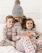 Smiling mom in a grey pom-pom beanie hugging her son and daughter on a bed wearing matching white Fair Isle reindeer pajama sets.