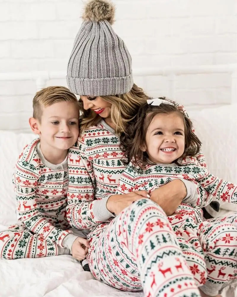 Smiling mom in a grey pom-pom beanie hugging her son and daughter on a bed wearing matching white Fair Isle reindeer pajama sets.