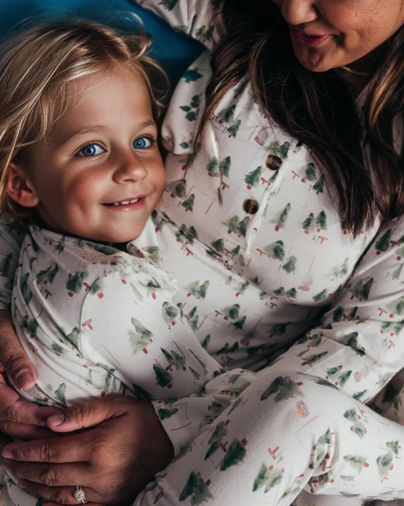 A close-up shot of a young blonde child hugging her mother, both wearing soft white matching holiday pajamas with green watercolor Christmas trees and henley button details.