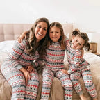 Happy mother sitting on a tufted headboard bed with her two daughters wearing matching white and red Fair Isle holiday pajama sets with grey cuffs.