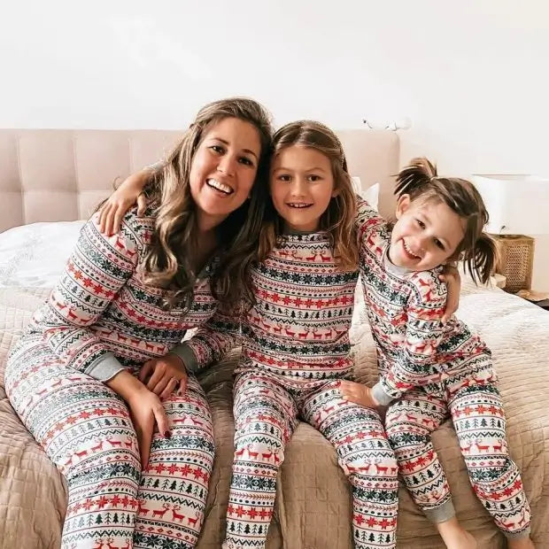 Happy mother sitting on a tufted headboard bed with her two daughters wearing matching white and red Fair Isle holiday pajama sets with grey cuffs.