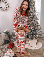 Mother in red and white Fair Isle pajamas holding a toddler wearing a matching onesie and reindeer hat next to a decorated Christmas tree.