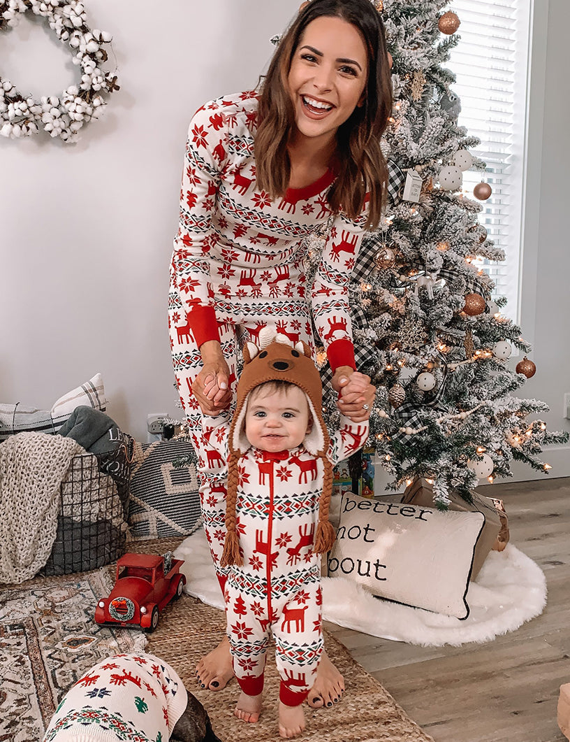 Mother in red and white Fair Isle pajamas holding a toddler wearing a matching onesie and reindeer hat next to a decorated Christmas tree.