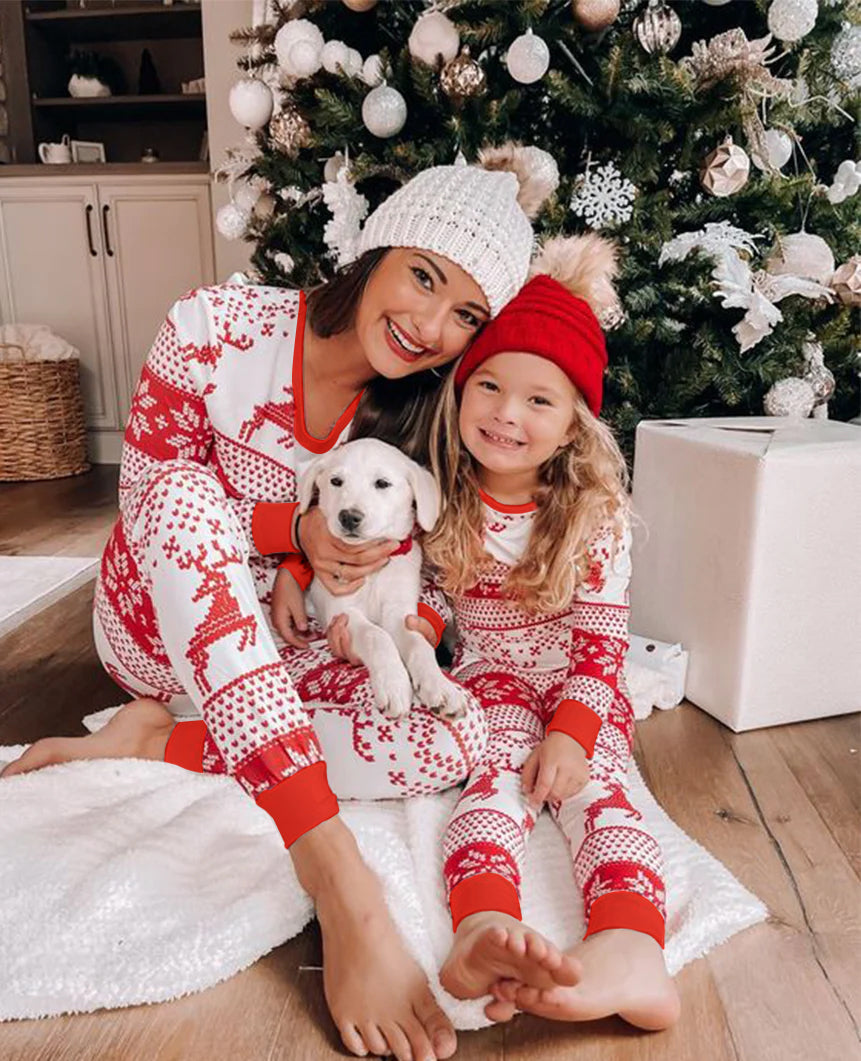 Mother and daughter sitting barefoot by a decorated Christmas tree wearing matching red beanies and red and white Fair Isle reindeer pajamas, holding a white puppy.