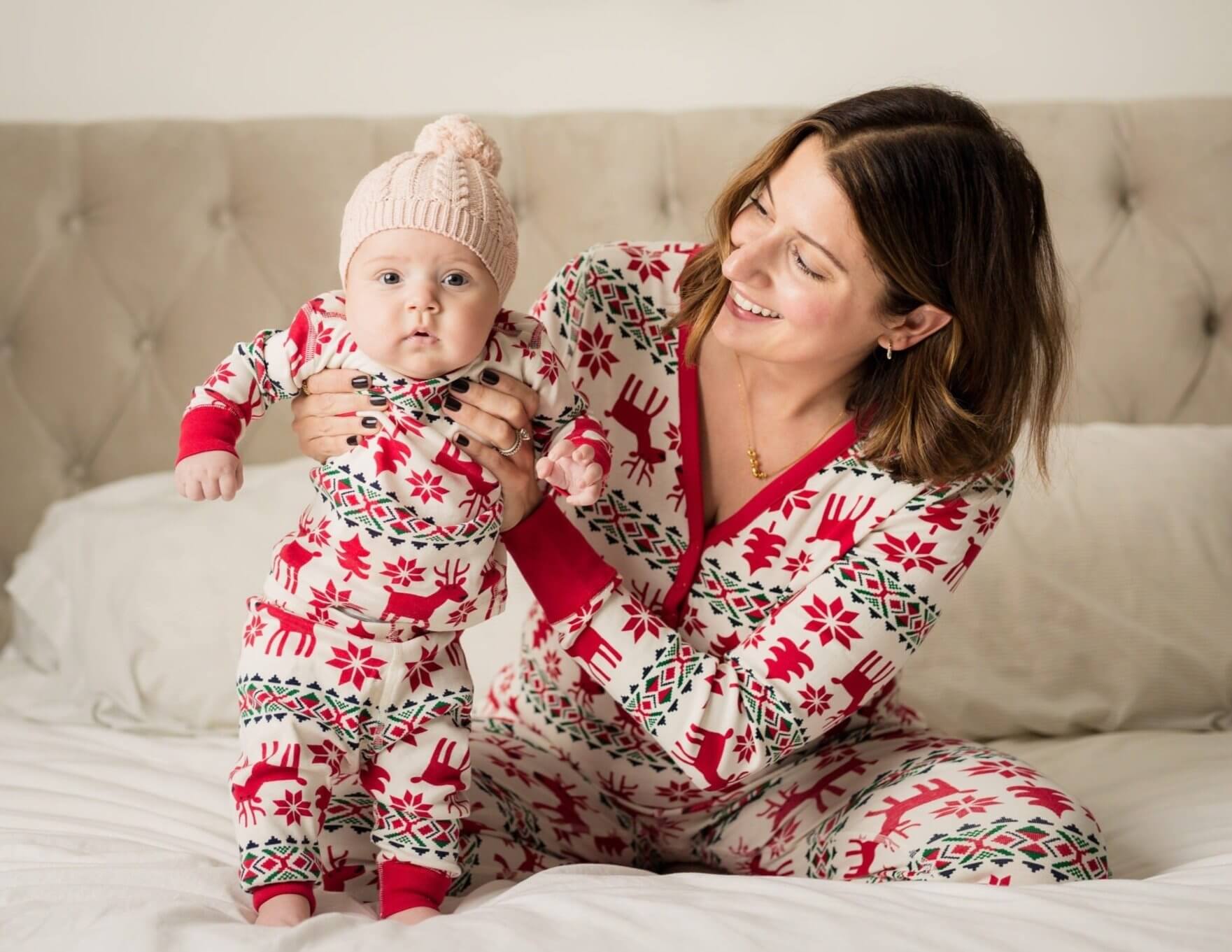 Mother holding an infant in matching red reindeer Fair Isle pajamas with a pink beanie while sitting on a tufted white bed.