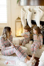 Mother sitting on a rug reading a book to her two toddlers by a fireplace with white Christmas stockings, all wearing matching Fair Isle pajamas.