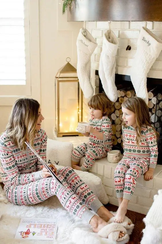 Mother sitting on a rug reading a book to her two toddlers by a fireplace with white Christmas stockings, all wearing matching Fair Isle pajamas.