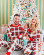 Smiling mother and father sitting in front of a pastel-decorated Christmas tree holding their crying newborn baby in a matching cream and red plaid onesie.