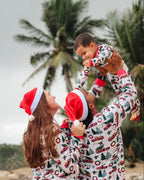 Father holding toddler high in the air beside mother under palm trees, wearing matching vintage red truck print Christmas pajamas and Santa hats.