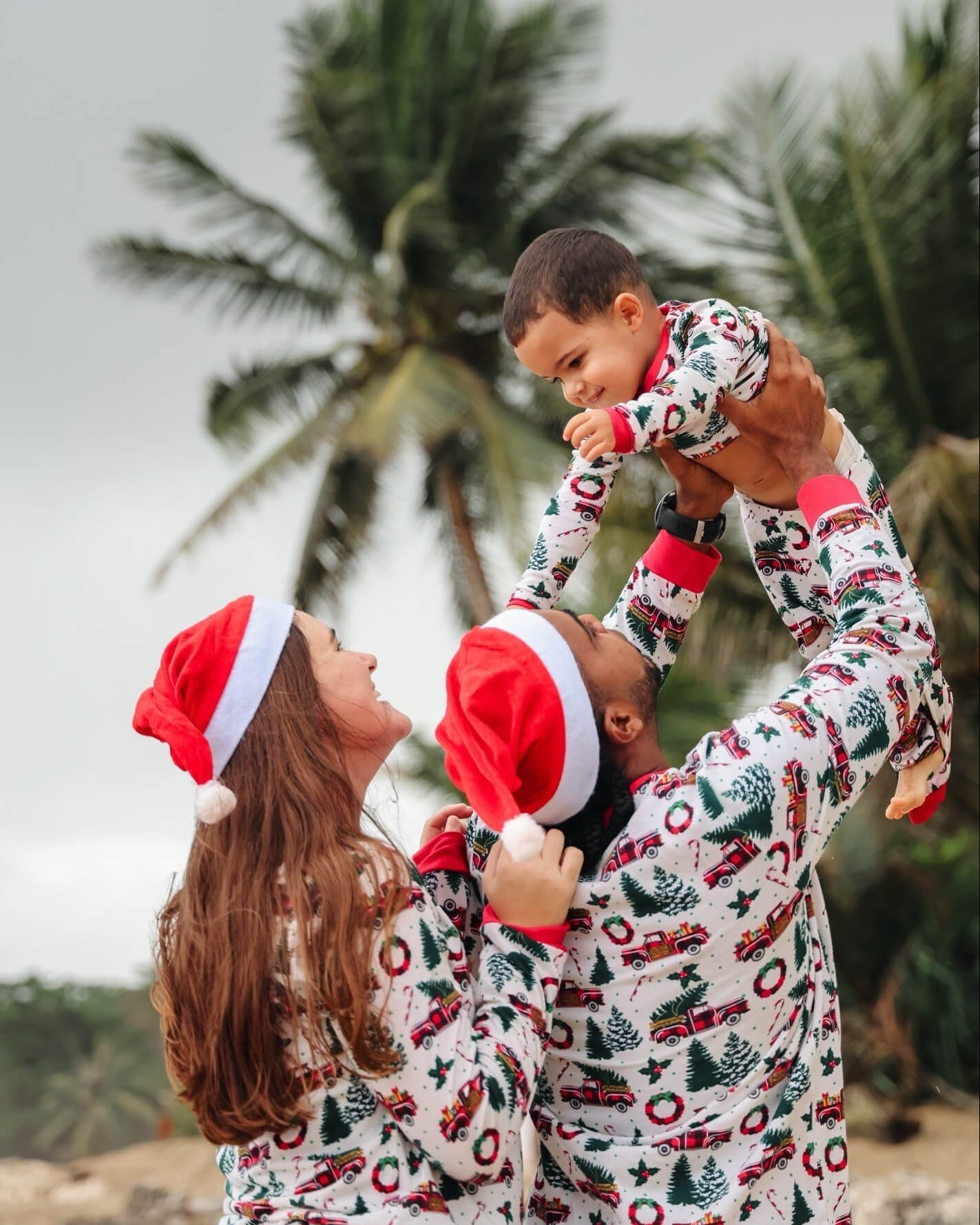 Father holding toddler high in the air beside mother under palm trees, wearing matching vintage red truck print Christmas pajamas and Santa hats.