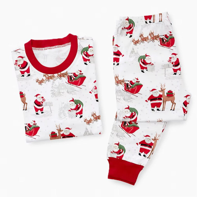 Flat lay of a white two-piece long-sleeve pajama set with red ribbed cuffs, printed with Santa Claus and reindeer pulling a sleigh.