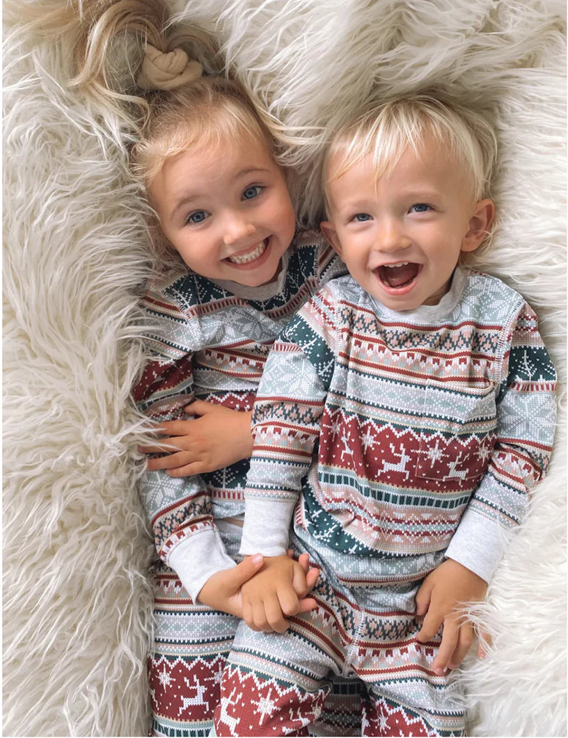 Two young children smiling on white faux fur rug wearing matching grey Fair Isle holiday PJs.