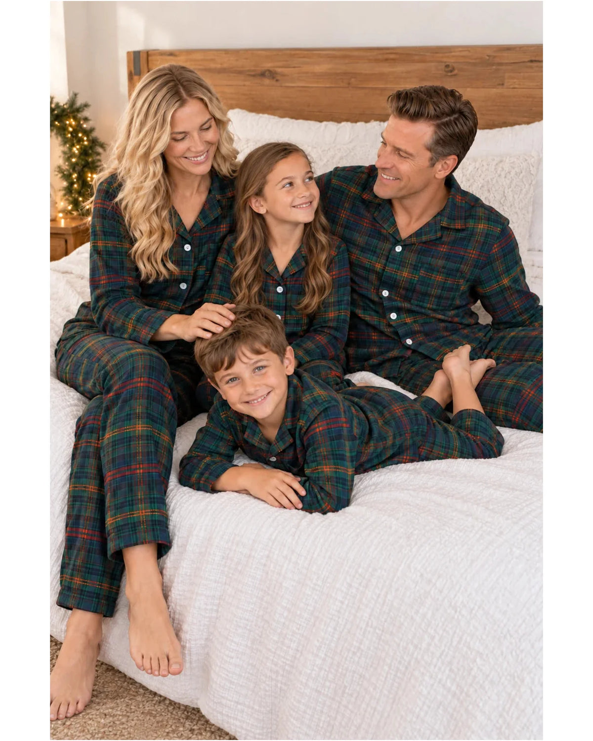 Family of four in dark green tartan pajamas sitting together on a bed.
