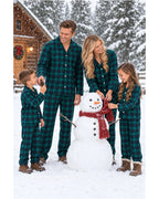 Family wearing green and black plaid pajamas beside a snowman outside a wooden cabin.