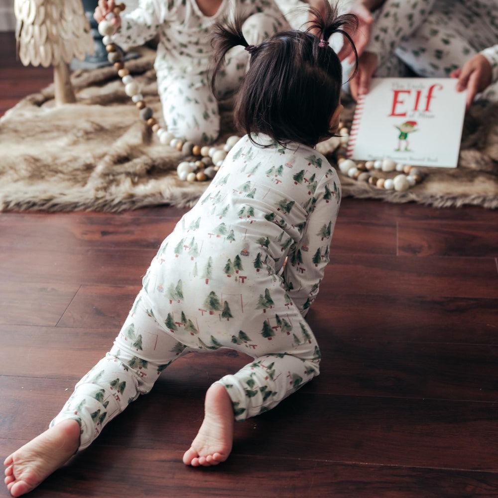 A toddler girl with pigtails crawling on a dark hardwood floor towards a Christmas book, wearing matching white pajama set with a watercolor evergreen tree print.