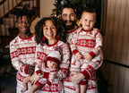 Happy toddler girl standing with hands on hips on a white fuzzy rug in front of a stone fireplace, modeling matching red and white Fair Isle reindeer kids Christmas pajamas.