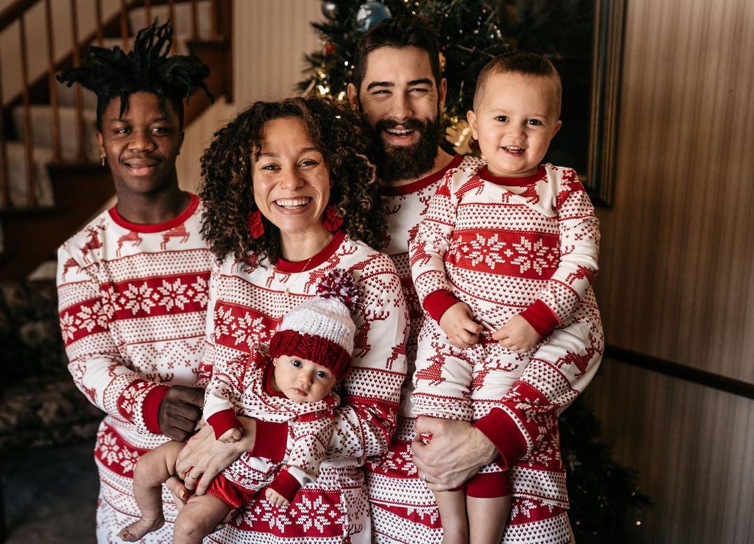 Happy toddler girl standing with hands on hips on a white fuzzy rug in front of a stone fireplace, modeling matching red and white Fair Isle reindeer kids Christmas pajamas.