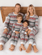 Family sitting against a wooden headboard in grey Christmas pajamas with red reindeer bands and green trees.