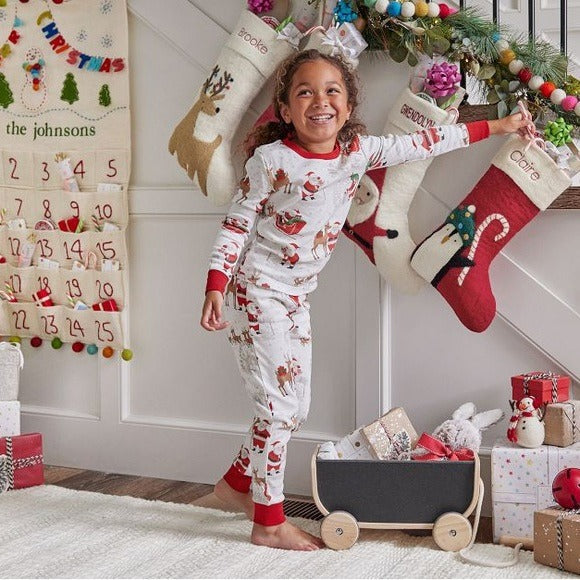 Smiling girl in long-sleeve white Santa print pajamas reaching for a stocking hanging on the wall near a Christmas countdown calendar.